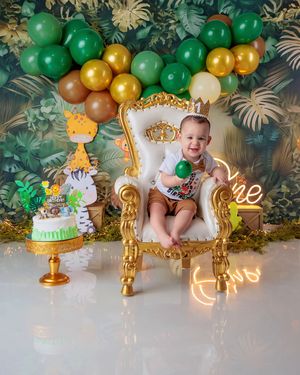 Smiling baby sits in golden throne chair at jungle safari first birthday celebration.