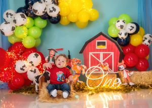 Baby sits in hay with farm themed backdrop featuring red barn, cow print balloons, and colorful green, red and yellow balloon arch.