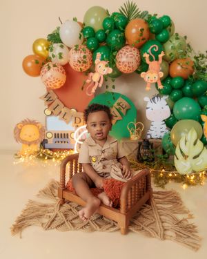 Baby boy in safari outfit sits on wooden bed with jungle animal balloon arch backdrop.