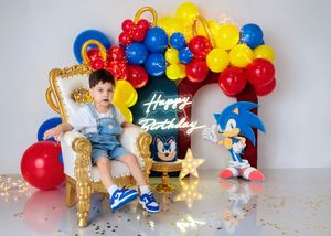 Young boy in denim overalls sits on gold throne with Sonic the Hedgehog themed red, blue and yellow balloon arch Happy Birthday setup.