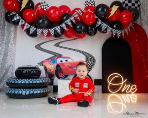 Baby boy in red racing suit at Cars themed first birthday with Lightning McQueen backdrop.
