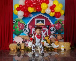 Baby sits before red barn farm birthday backdrop with balloon arch and hay bales.