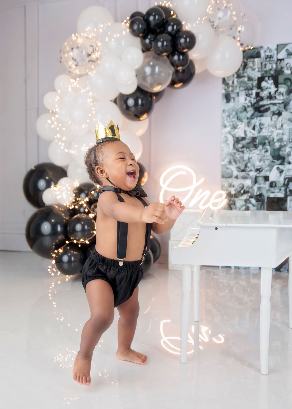 A laughing baby boy in black suspenders and a gold crown stands before a black and white balloon arch with neon One sign.