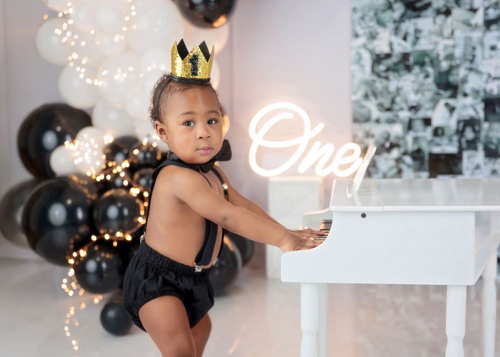 A baby boy in black suspenders and gold crown leans on a white mini piano beside a black and white balloon garland.