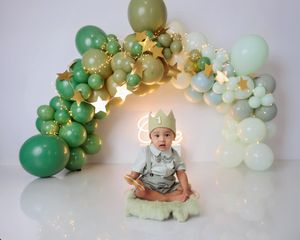 Baby in crown sitting before green and gold balloon arch at first birthday photo session.