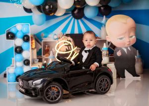 Baby boy in tuxedo sits in toy Mercedes car with Boss Baby themed blue and black balloon decorations.