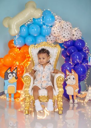 Baby sits on ornate gold throne with Bluey themed balloon arch featuring orange, blue and white balloons with dog bone topper.