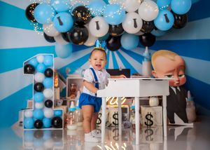 Baby boy in blue outfit plays piano with Boss Baby themed blue, black, and white balloon decorations.