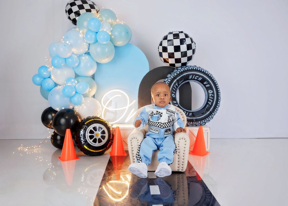 A baby boy sits in a small chair before a racing-themed backdrop with checkered balloons, tires, and orange traffic cones.