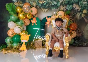 A toddler boy sits on a golden throne chair at a safari jungle themed second birthday party with giraffe decorations.
