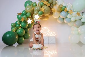 Baby smiling at first birthday cake smash with green and gold balloon arch and 'One' neon sign.