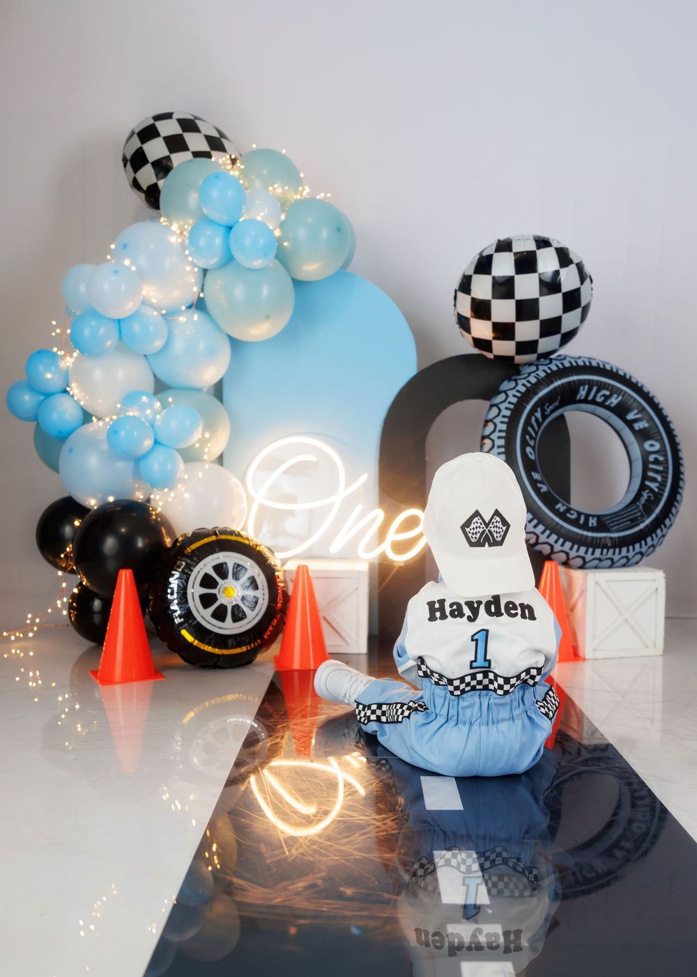 A baby named Hayden sits before a racing-themed cake smash setup with checkered balloons, tires, and orange cones.
