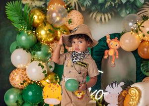 Toddler in khaki safari outfit stands among colorful jungle balloons and animal decorations at birthday party.