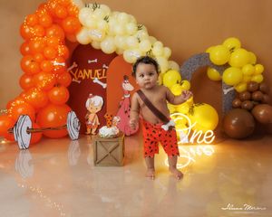 Baby boy in Flintstones themed first birthday cake smash with orange balloons and colorful decorations.