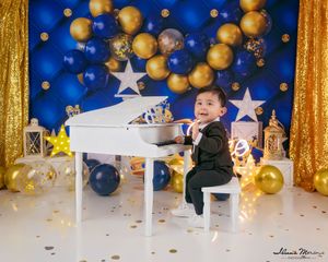 Smiling toddler boy in tuxedo sitting at white toy piano with blue and gold star decorations.