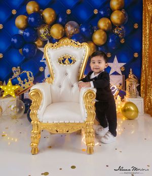 Toddler boy in tuxedo standing beside ornate gold and white throne with blue and gold balloons.