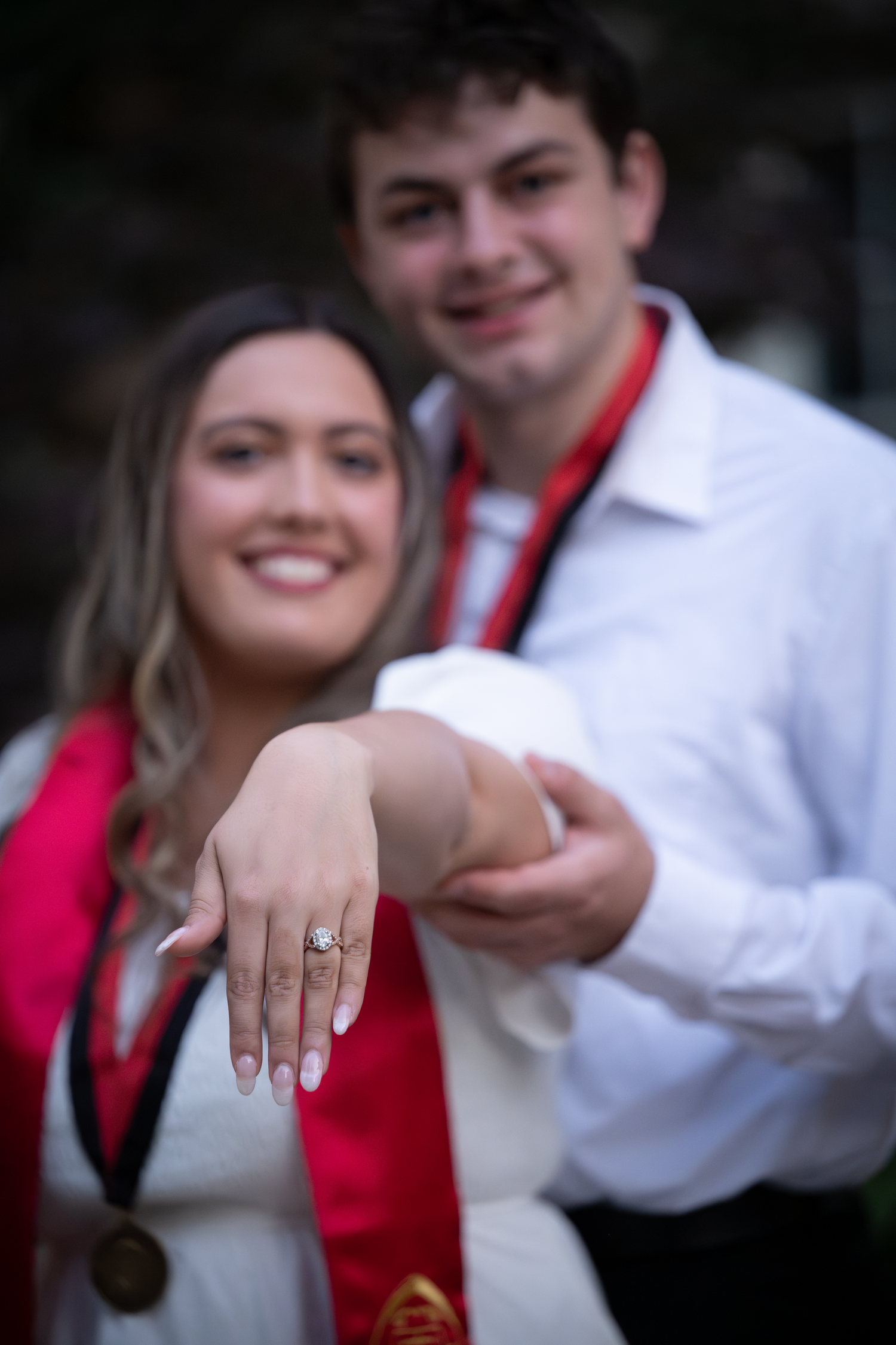 Couple proudly displaying engagement ring, wearing graduation sashes and medal, smiling joyfully at their celebration.