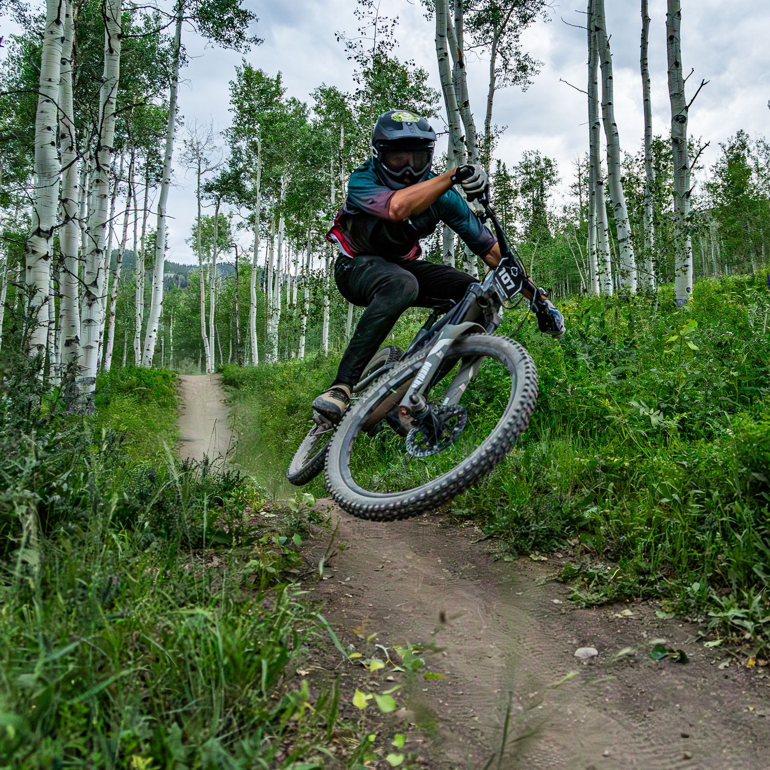 Mountain biker performs aerial jump on dirt trail through aspen forest wearing full protective gear on sunny day.