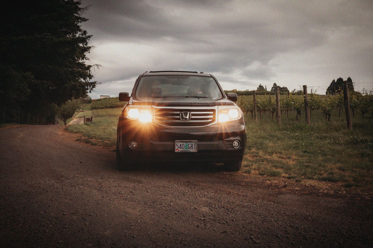 Dark Honda Pilot SUV with headlights on driving along a rural dirt road beside a vineyard under cloudy skies.
