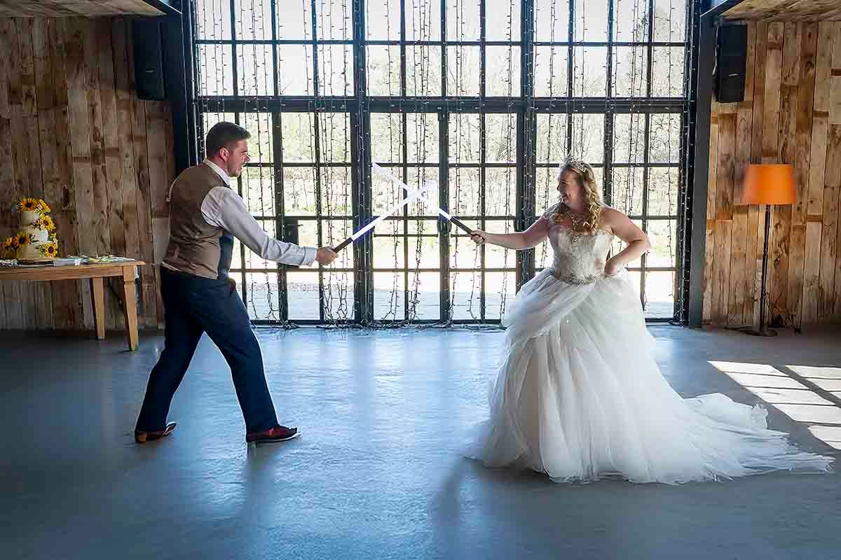 Bride in white tulle gown and groom in vest playfully duel with swords during wedding reception in rustic barn venue.