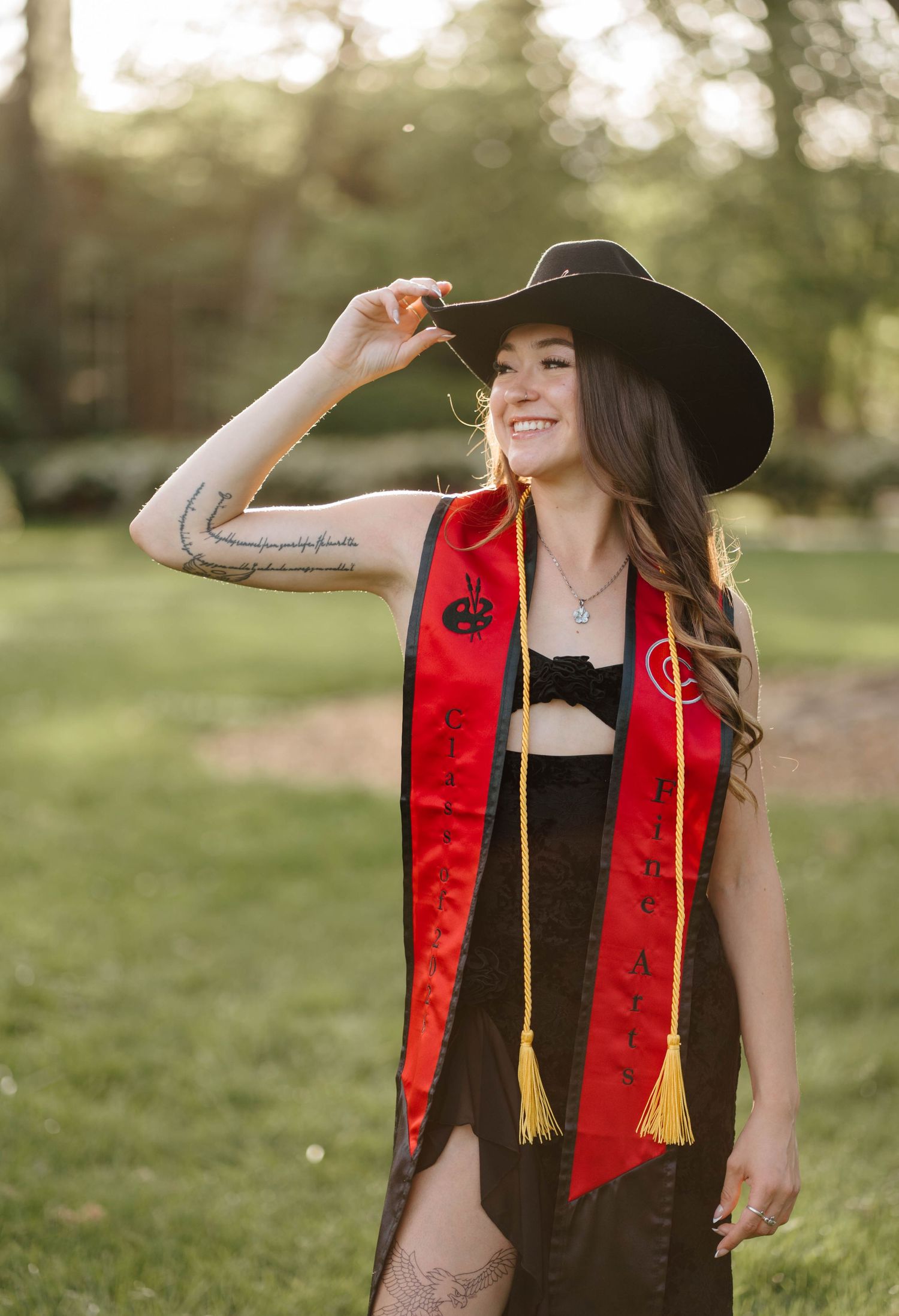Smiling graduate woman wearing red sash and black cowboy hat poses outdoors in park setting.