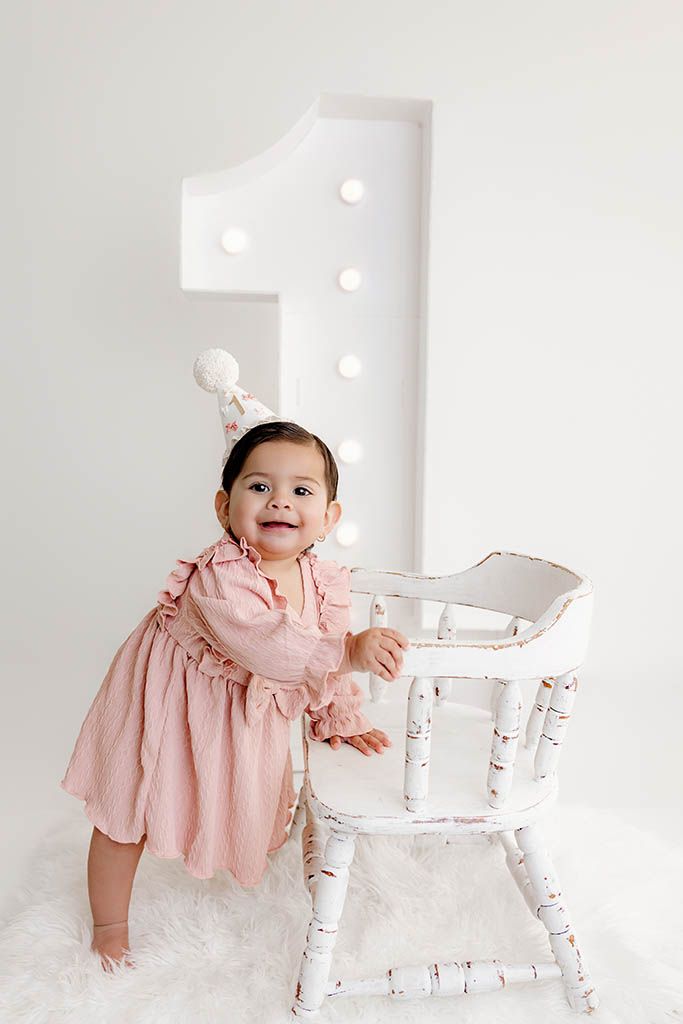 Smiling baby girl in pink dress standing by white chair with large illuminated number one backdrop for first birthday.