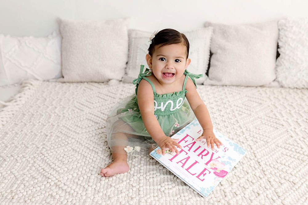 Baby girl in green one romper sits on bed holding Fairy Tale book.