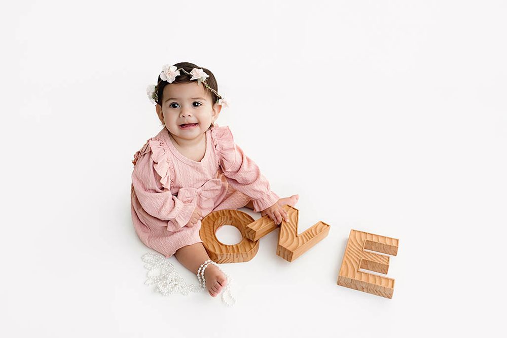 Baby girl in pink ruffled dress sits with wooden LOVE letters on white background.