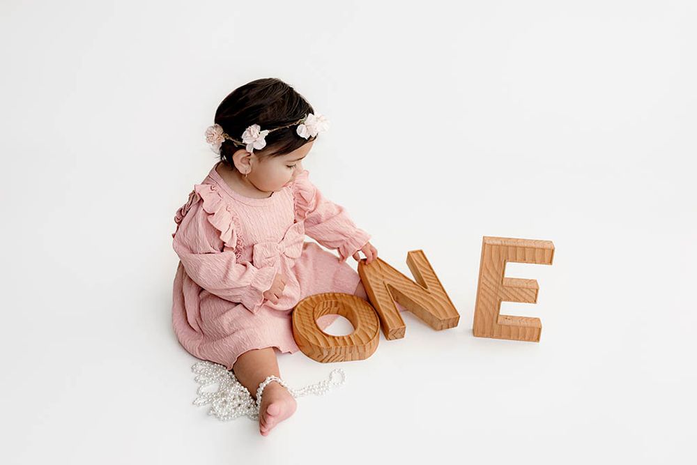 Baby girl in pink dress sits beside wooden ONE letters celebrating her first birthday photo shoot.