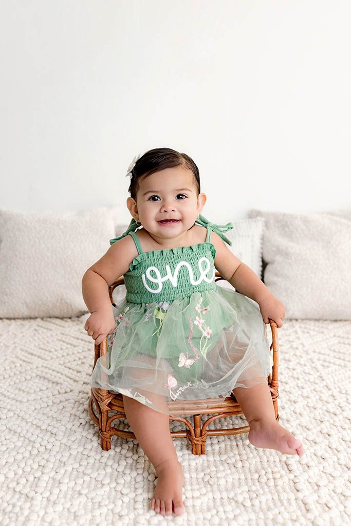 Happy baby girl in green tulle one dress sitting in rattan chair for first birthday photo session.