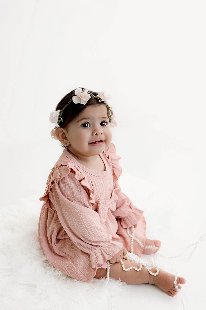 Baby girl in pink ruffle romper and floral crown sits on white fur celebrating her 1st birthday.