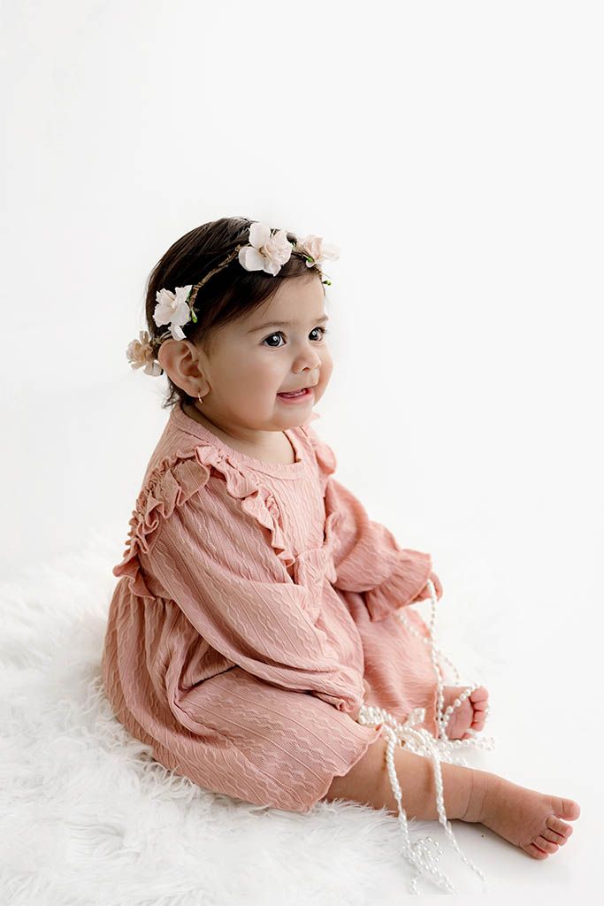 Smiling baby girl in pink ruffle romper and floral crown poses on white fur rug.