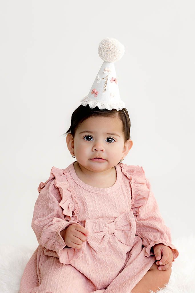 Adorable baby girl in pink ruffle outfit and white birthday party hat by Menifee first birthday photographer.