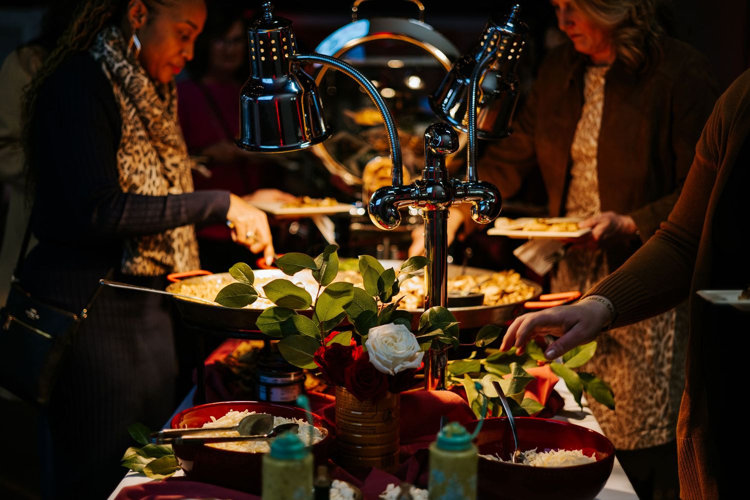 Guests serving themselves at a festive buffet table with warm food under heat lamps and floral centerpiece.