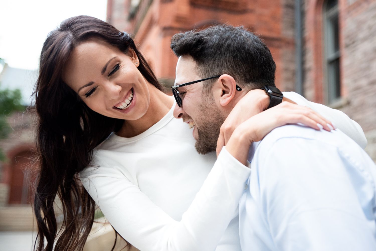 A smiling couple sharing a joyful moment outdoors, woman in white top embracing man in sunglasses.
