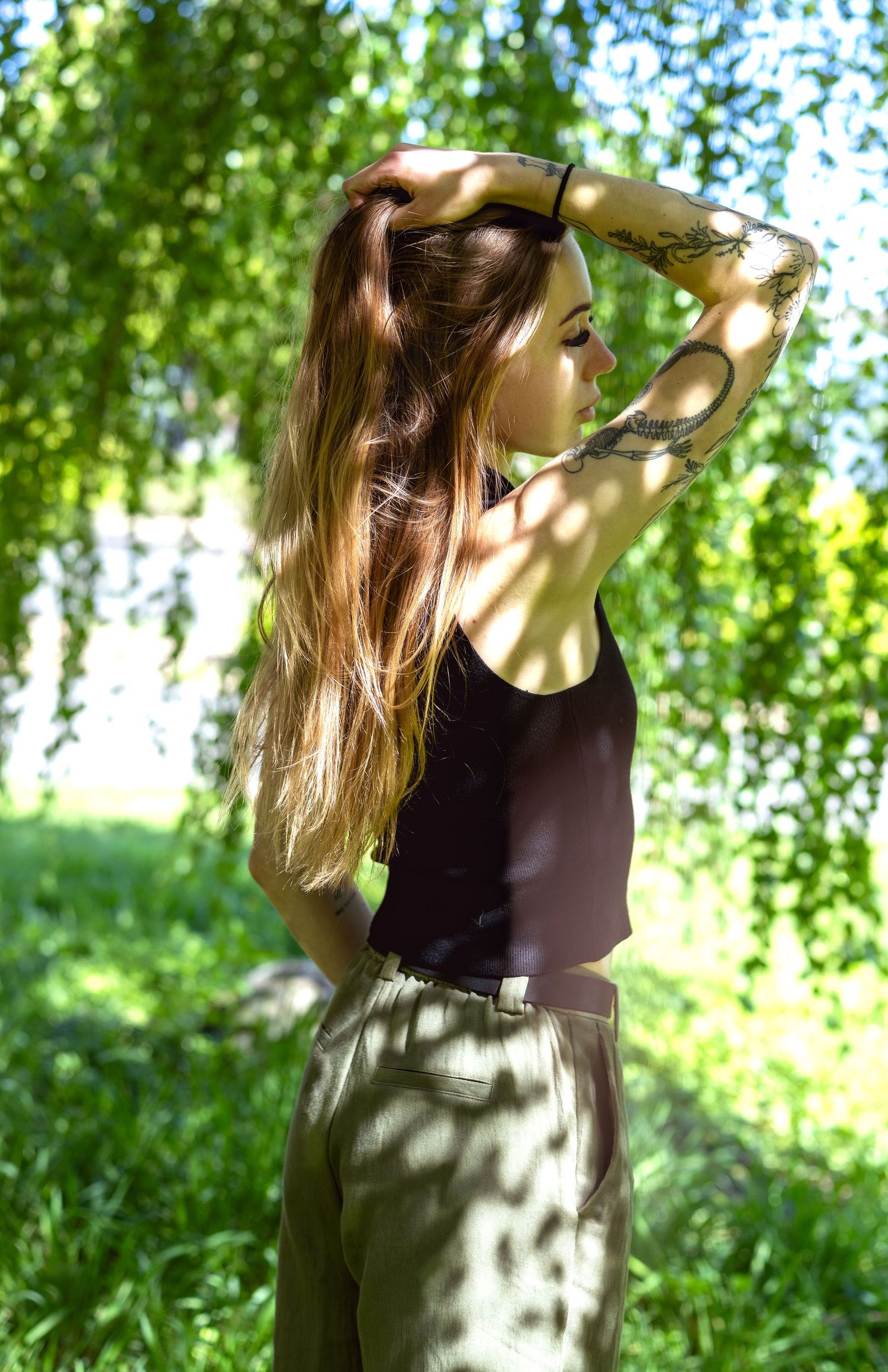 Side portrait of a person with tattoos standing in dappled sunlight under green trees.