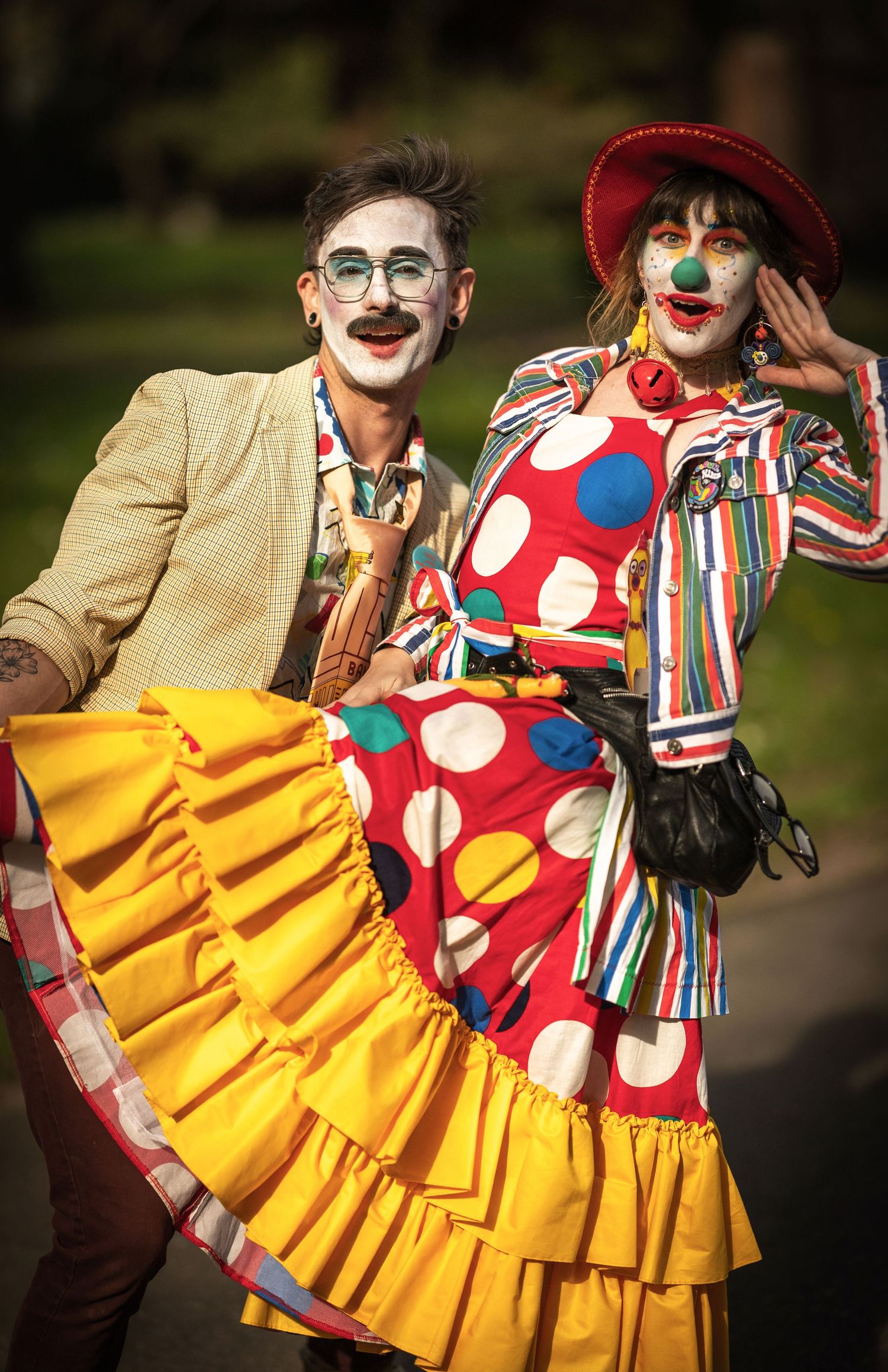 Two clowns in colorful costumes pose together outdoors in warm sunlight.