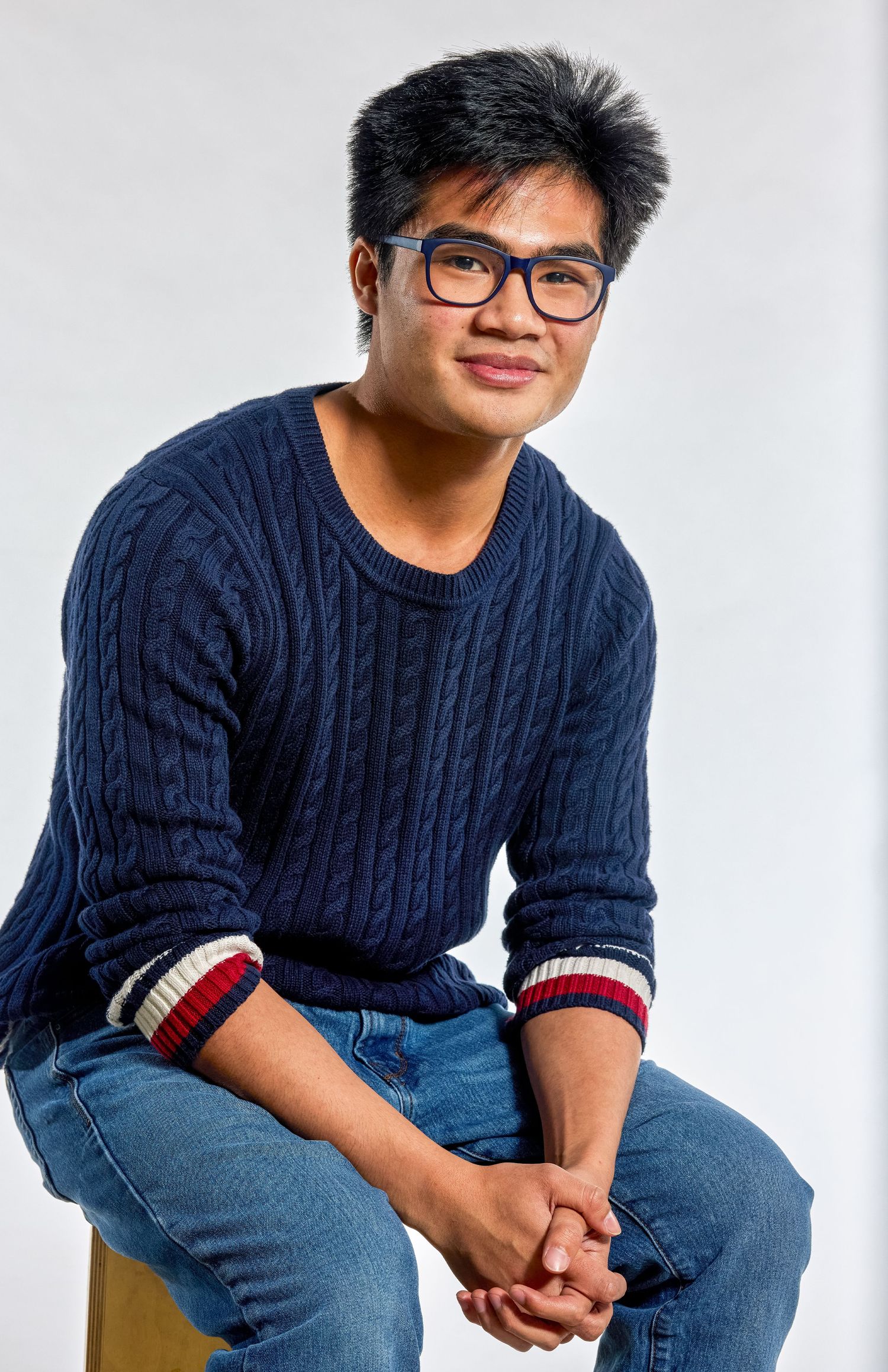 Seated studio portrait of a man wearing glasses and a navy sweater against a light background.