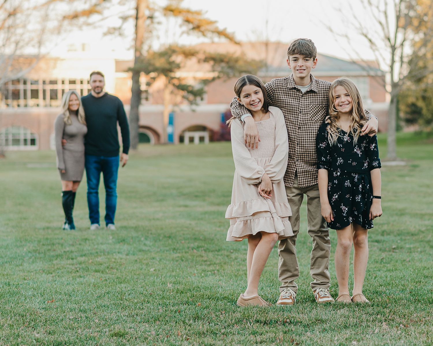 Family portrait with two parents standing behind three children posing on a green lawn near a brick building.