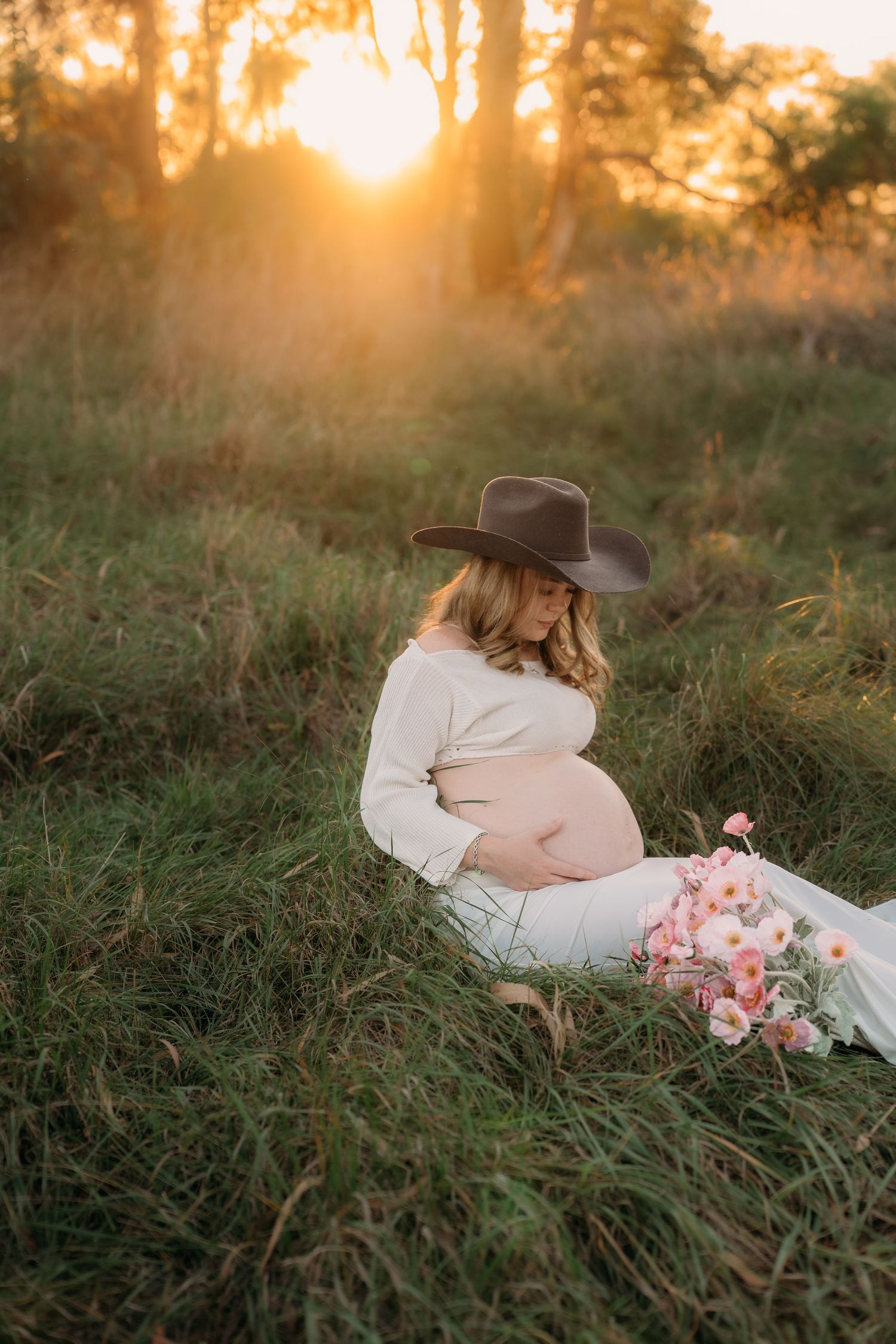 Pregnant woman in cowboy hat sits in golden sunset field with pink flowers, cradling bare baby bump in white dress.