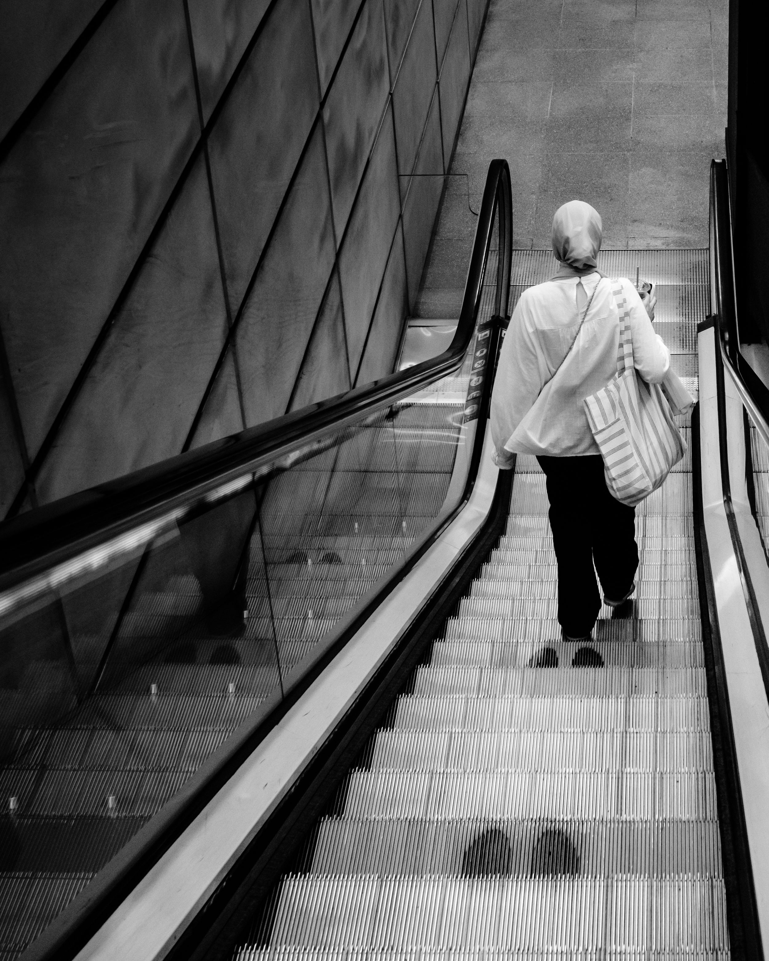 Black and white photo of an elderly person riding an escalator inside a modern glass structure