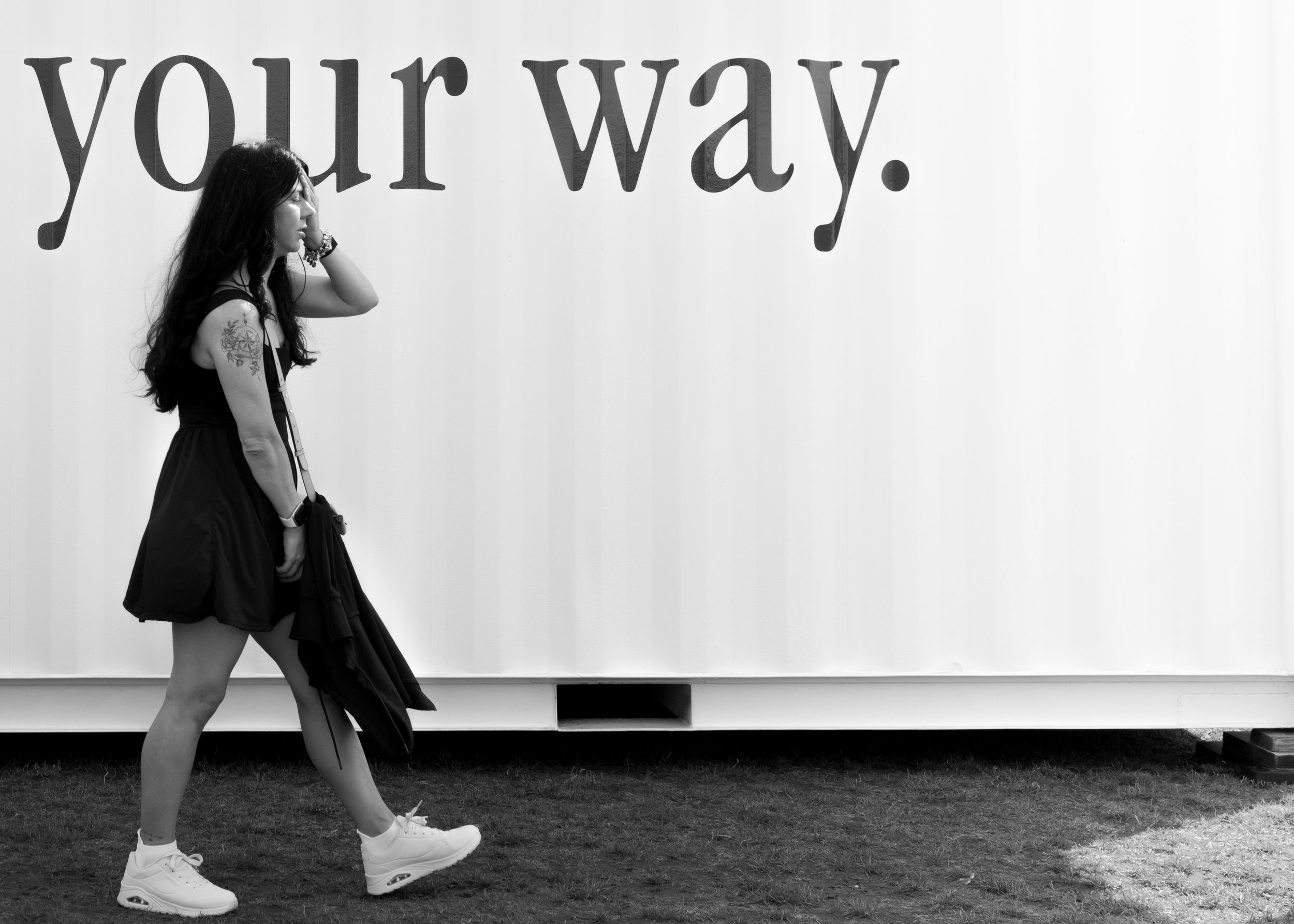 Black and white street photography of a woman walking past a large sign reading your way