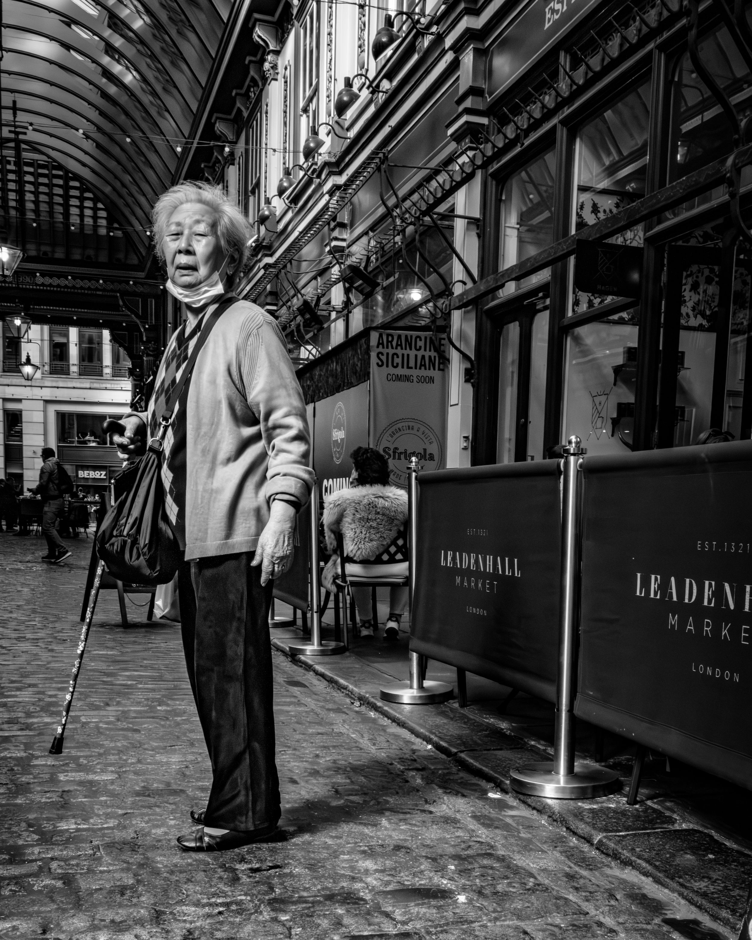 Black and white street photography of an elderly man walking with a cane through a market arcade