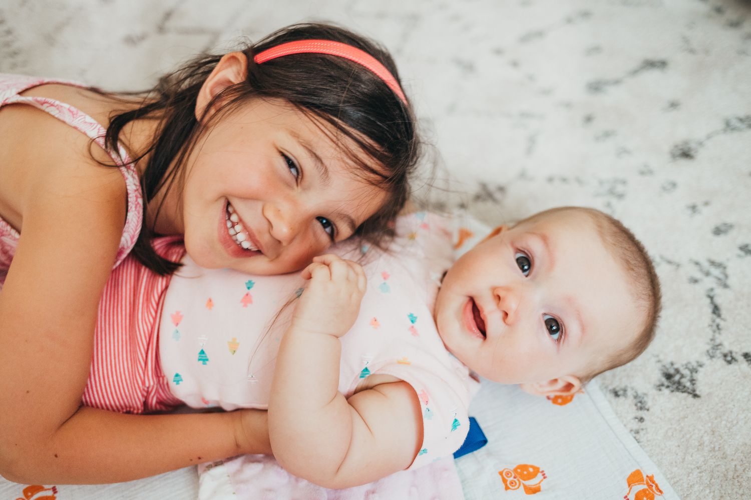 Smiling young girl lying next to a happy baby on a soft white blanket, siblings sharing a joyful moment.