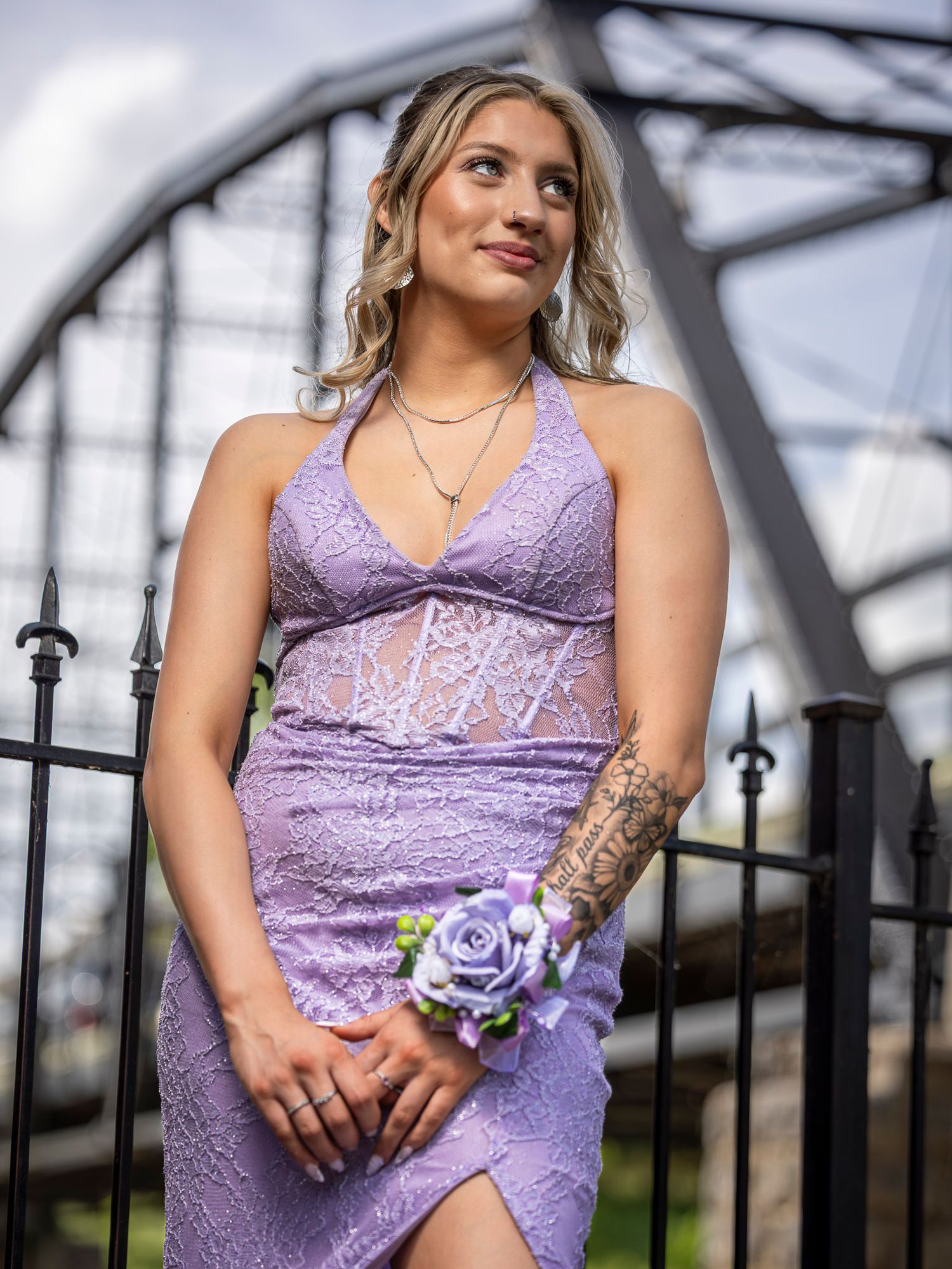 Young woman in lavender lace halter prom dress with floral wrist corsage poses near decorative bridge.