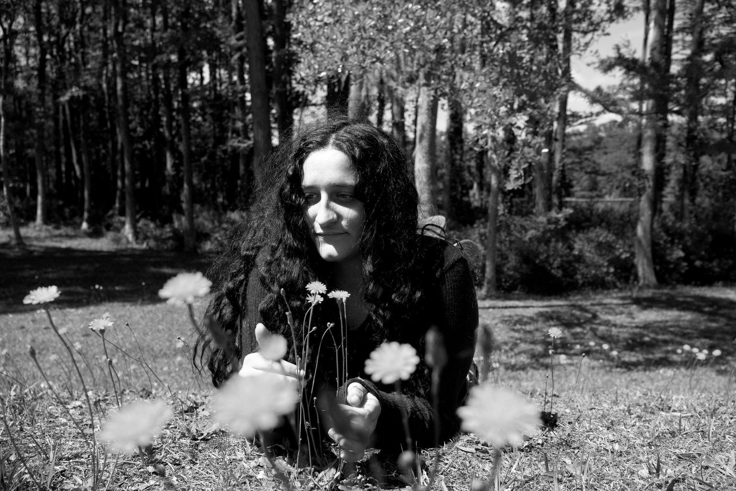Woman with dark curly hair crouching among yellow dandelion flowers in a sunny green forest clearing.