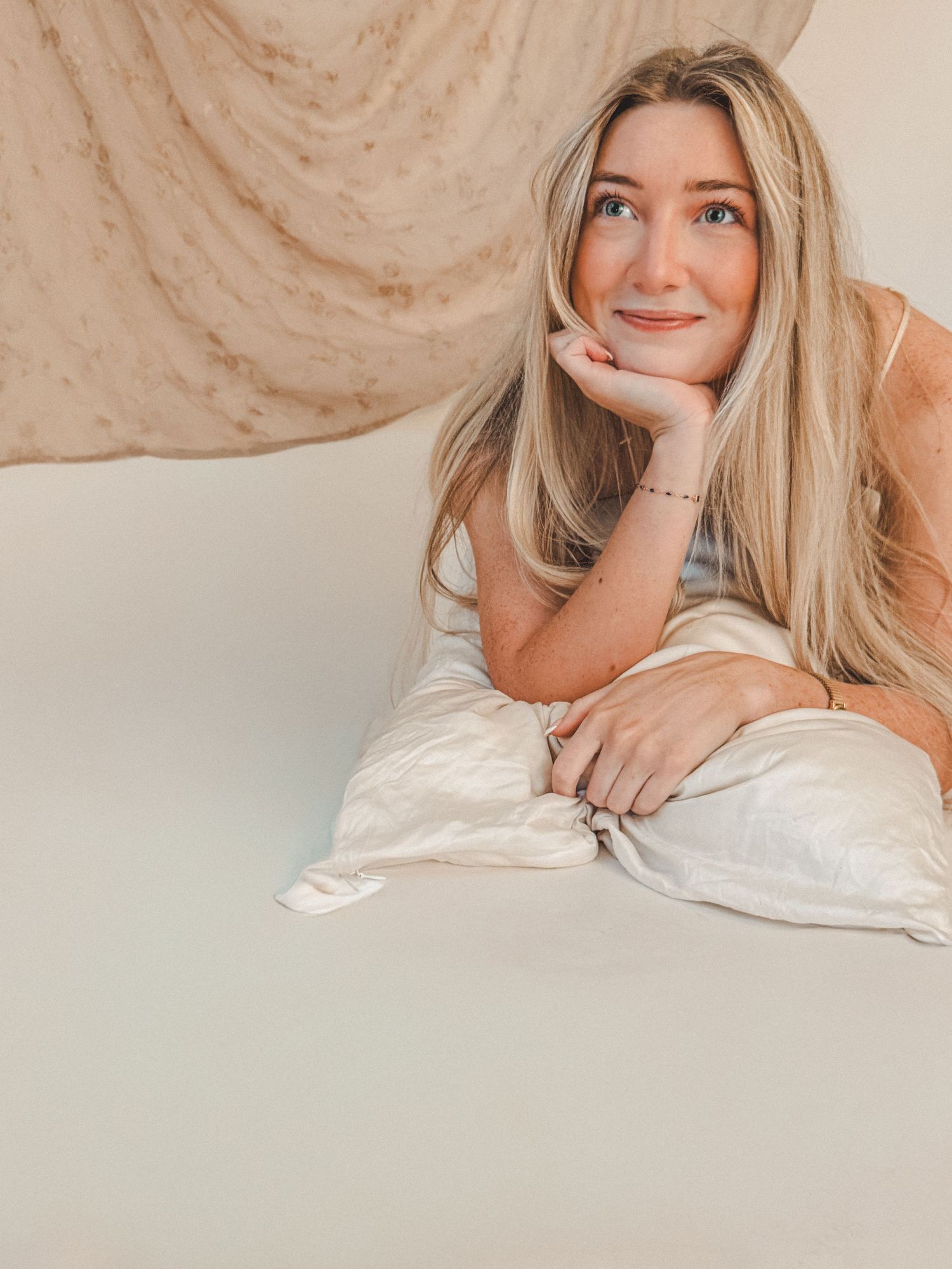 Smiling blonde woman with blue eyes resting chin on hand while lying on white pillow with neutral backdrop.