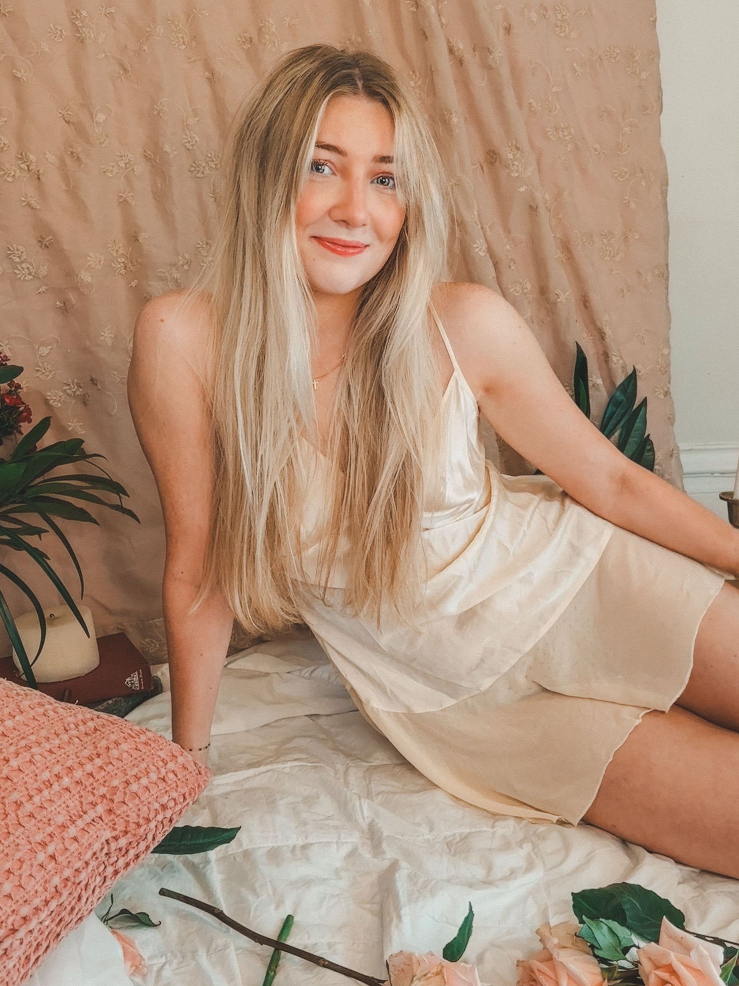 Smiling blonde woman in a cream satin slip dress sitting on floral bedding with a pink backdrop.
