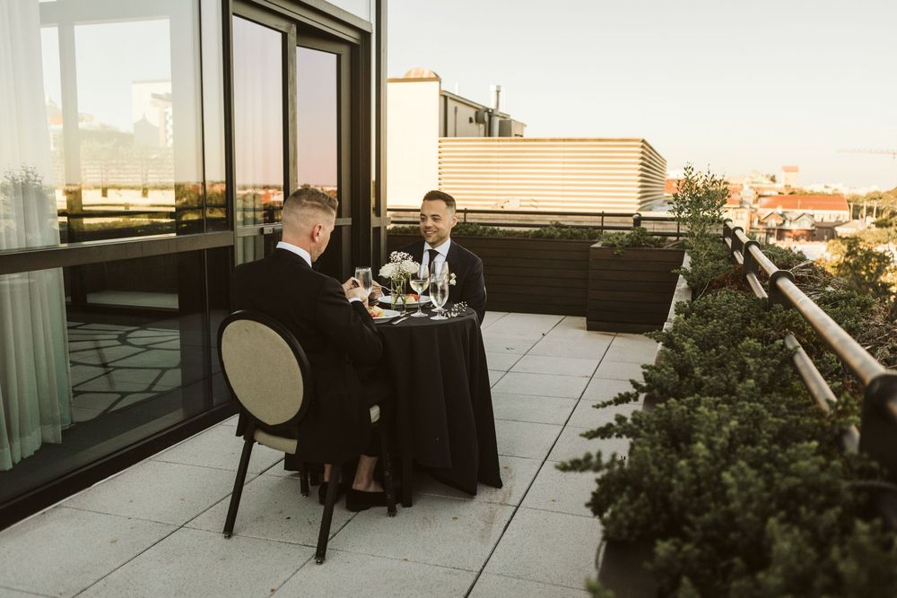 Two men in formal attire share an intimate rooftop dinner at sunset with city views.