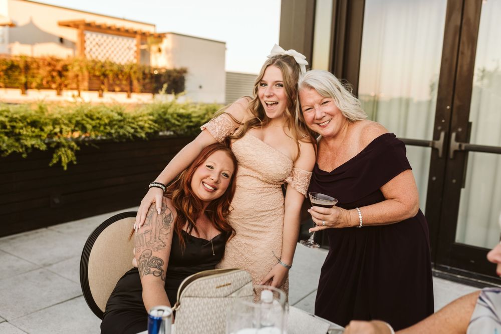 Three women smiling and laughing together at an outdoor rooftop event, one wearing a bridal veil.